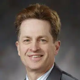 Adam Reynolds is a white middle-aged male wearing a dark shiny jacket, with pinstriped collared shirt and blue tie. He has short brown hair and is smiling at you.