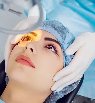 Cataracts: A female patient wearing a blue surgical cap looks up into a light shining into her right eye. A pair of hands wearing white surgical gloves hold her head behind her.