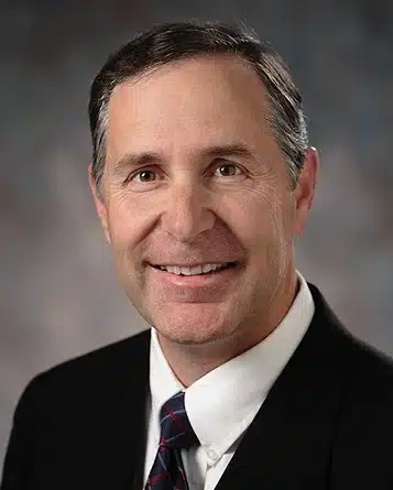 An older white man with short dark hair, graying at the temples, and wearing a black suit with white shirt and dark tie, smiles at you.