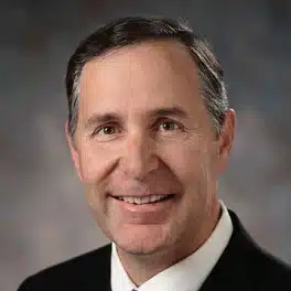 An older white man with short dark hair, graying at the temples, and wearing a black suit with white shirt and dark tie, smiles at you.