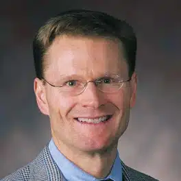A middle aged white man with short brown hair, rimless glasses, is wearing a gray glencheck jacket, blue denim shirt, and dark reddish gray tie. He is smiling at you.