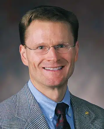 A middle aged white man with short brown hair, rimless glasses, is wearing a gray glencheck jacket, blue denim shirt, and dark reddish gray tie. He is smiling at you.