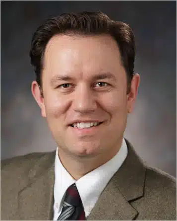 Lee Carter has short brown hair, a dimple on his chin, and is wearing a brown jacket with white collar and striped red-gray tie. He smiles at you.