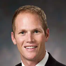 A tall white man with blue eyes, short blonde hair, black jacket, white shirt, and red tie is smiling at you. He looks tall.