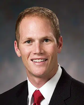 A tall white man with blue eyes, short blonde hair, black jacket, white shirt, and red tie is smiling at you. He looks tall.