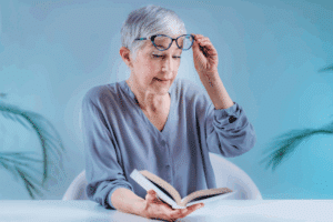 An older woman with glasses reads a book, interrupted by her cataract symptoms and needs treatment information.