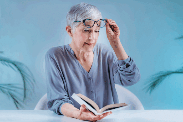 An older woman with glasses reads a book, interrupted by her cataract symptoms and needs treatment information.