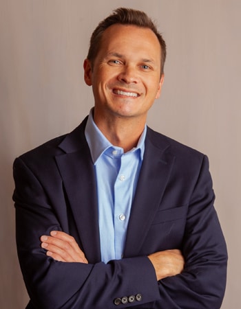 A young white man with short brown hair is wearing a dark blue coat over a light blue collard shirt, and smiling at you.